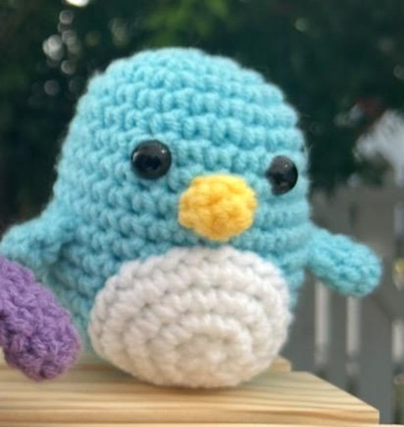 May include: A crocheted bird toy, featuring a light blue body, white belly, and yellow beak. It has black safety eyes and a small purple wing. This soft toy is ideal for a child's room or as a decorative item.