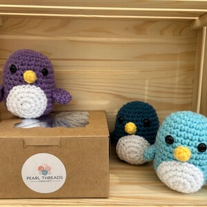 May include: Handmade crochet penguin toys in purple, teal, and light blue with white bellies and yellow beaks. The toys are displayed in a wooden crate with a cardboard box featuring a "Pearl Threads Crochet" sticker.