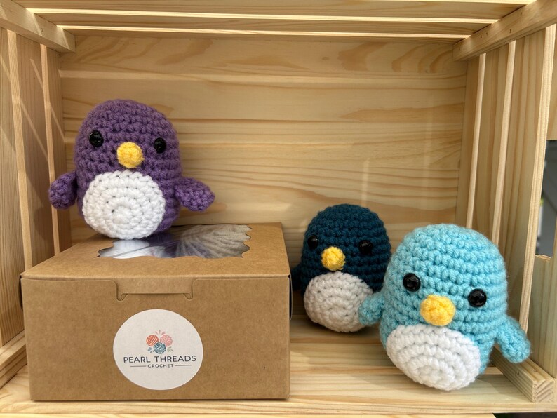 May include: Handmade crochet penguin toys in purple, teal, and light blue with white bellies and yellow beaks. The toys are displayed in a wooden crate with a cardboard box featuring a "Pearl Threads Crochet" sticker.