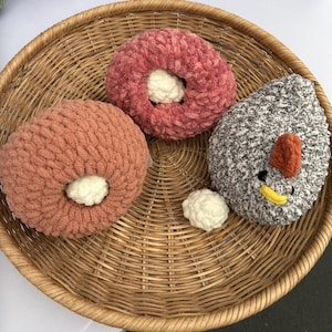 May include: A wicker basket contains three handmade plush toys. Two are donut-shaped, in pink and peach with white centres. The third is a chicken, grey and white with an orange comb and yellow beak. A small white ball is also in the basket.