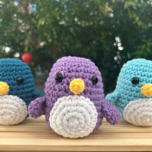May include: Three handmade crochet penguins in teal, purple, and light blue. Each penguin has a white belly, yellow beak, and black eyes. They are displayed on a wooden surface with a blurred green background.