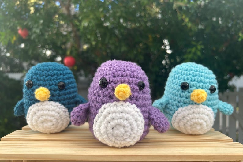May include: Three handmade crochet penguins in teal, purple, and light blue. Each penguin has a white belly, yellow beak, and black eyes. They are displayed on a wooden surface with a blurred green background.