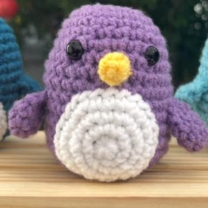 May include: A handmade crochet penguin in shades of purple, white, and yellow. The penguin has black button eyes and a yellow beak. The body is a soft, textured yarn, and the penguin is sitting on a wooden surface.