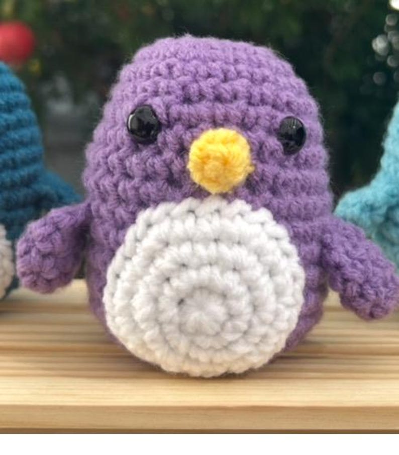 May include: A handmade crochet penguin in shades of purple, white, and yellow. The penguin has black button eyes and a yellow beak. The body is a soft, textured yarn, and the penguin is sitting on a wooden surface.