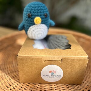 May include: A teal, white, and yellow crochet penguin sits on a brown cardboard box. The box has a circular sticker with the text "Pearl Threads Crochet". The penguin is on a woven basket.