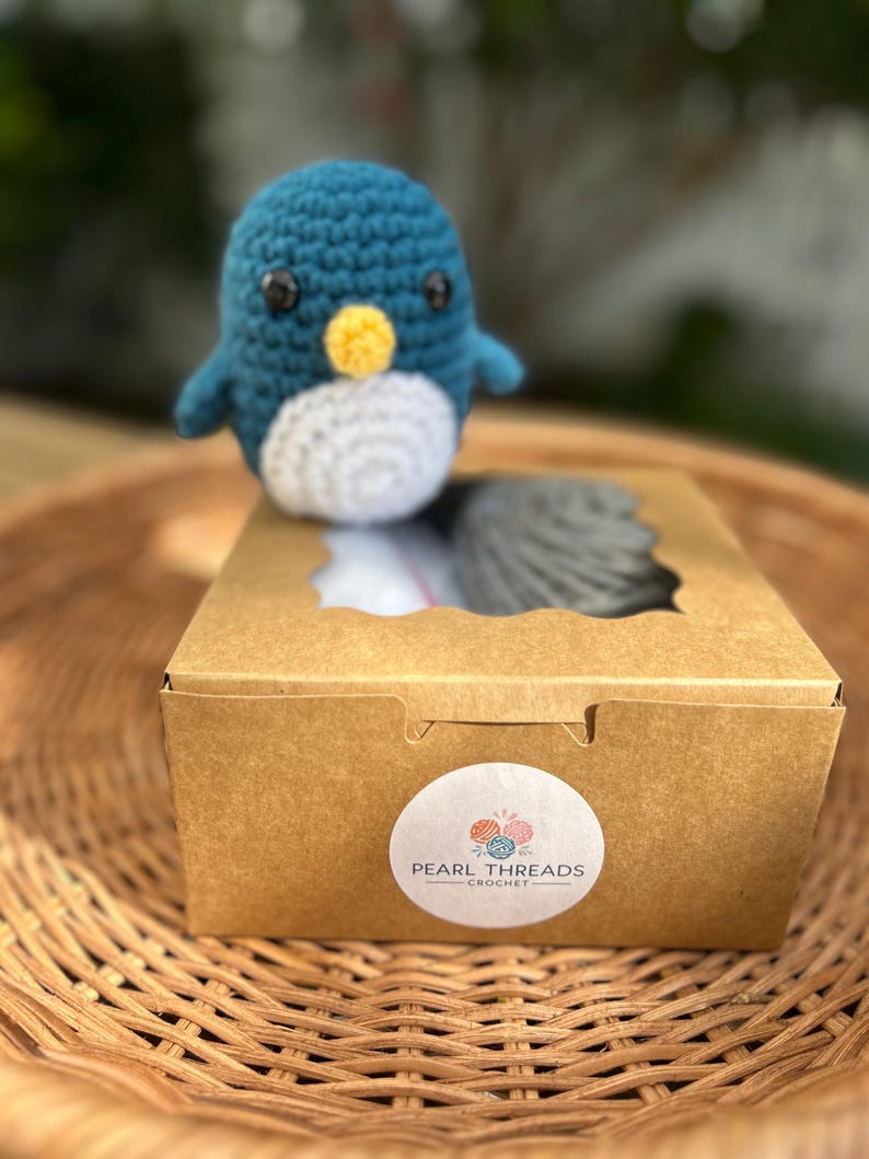 May include: A teal, white, and yellow crochet penguin sits on a brown cardboard box. The box has a circular sticker with the text "Pearl Threads Crochet". The penguin is on a woven basket.