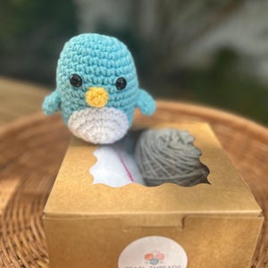 May include: A crocheted penguin toy in shades of blue, white, and yellow, with black eyes. It rests on a brown cardboard box containing grey yarn and a pink item. The box has a clear window and a "Pearl Threads Crochet" sticker.