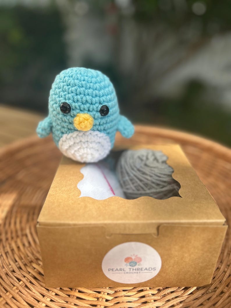 May include: A crocheted penguin toy in shades of blue, white, and yellow, with black eyes. It rests on a brown cardboard box containing grey yarn and a pink item. The box has a clear window and a "Pearl Threads Crochet" sticker.