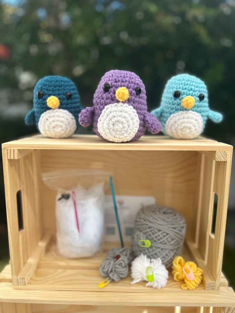 May include: Three crocheted penguin toys in teal, purple, and light blue, each with a white belly and yellow beak, sit on a wooden crate. The crate contains yarn, a crochet hook, and other crafting supplies, suggesting a DIY craft kit.