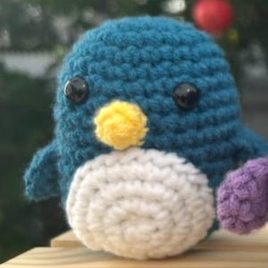May include: A teal blue, white, yellow, and purple crochet penguin. The penguin has black safety eyes and a yellow beak. The penguin is handmade. It is sitting on a wooden surface. The background is blurred with a red bauble.