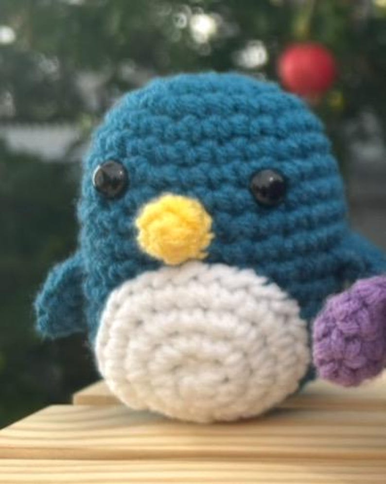 May include: A teal blue, white, yellow, and purple crochet penguin. The penguin has black safety eyes and a yellow beak. The penguin is handmade. It is sitting on a wooden surface. The background is blurred with a red bauble.