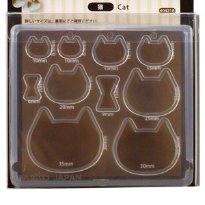 Puede incluir: Un molde de plástico transparente con nueve moldes de diferentes formas de gato. Los moldes están etiquetados con su tamaño en milímetros. El molde está etiquetado como "Soft Mold Cat" y está hecho por PADICO.