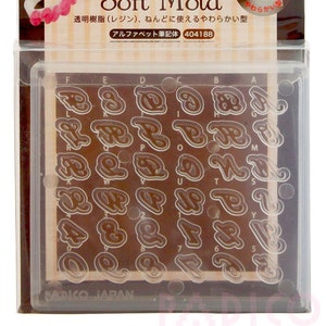 Puede incluir: Un molde de plástico transparente con un fondo marrón que contiene letras mayúsculas y números en una fuente cursiva. El molde está etiquetado como "Soft Mold" y "Alphabets in cursive".