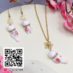 May include: A set of delicate gold jewelry featuring a white ceramic cat charm with pink floral accents. The necklace has a delicate gold chain and a small flower charm. The earrings are drop earrings with a flower charm and a cat charm.