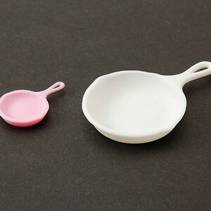 May include: Two miniature frying pans, one pink and one white, on a dark gray surface.