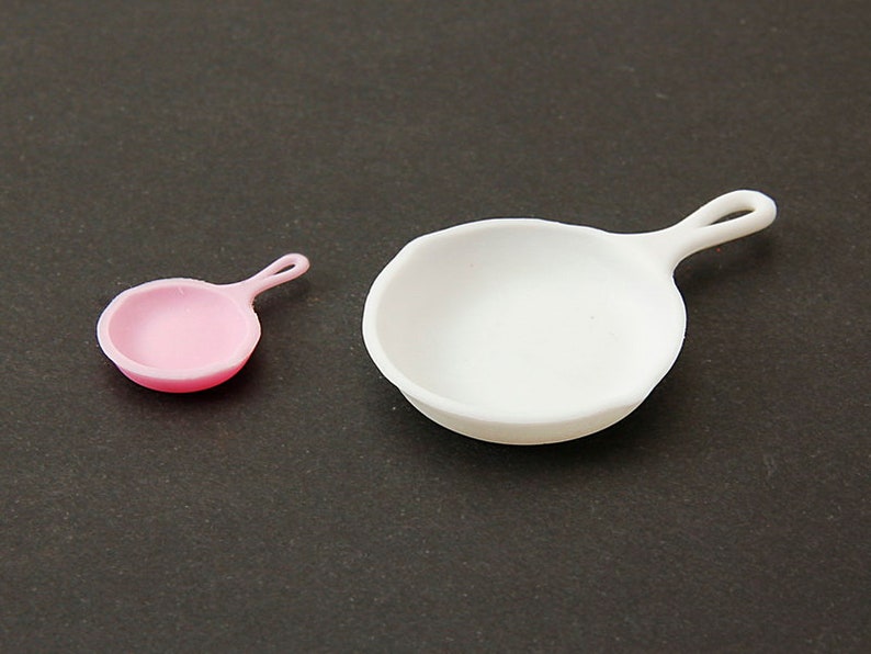 May include: Two miniature frying pans, one pink and one white, on a dark gray surface.