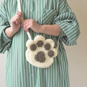 May include: A cream-colored, crocheted paw-shaped crossbody bag with brown paw pad details. The bag has a long, cream-colored strap and is held by a person wearing a green and white striped shirt.