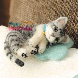 May include: A needle felted grey and white tabby cat with blue eyes, wearing a blue collar with a gold bell, lying on a light blue felt cushion.