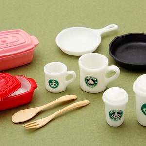 May include: A set of miniature kitchen and coffee shop items, including a pink and red casserole dish, a black skillet, a white frying pan, two white mugs with green logos, and two white to-go coffee cups with green logos. There are also two wooden utensils, a spoon and a fork.