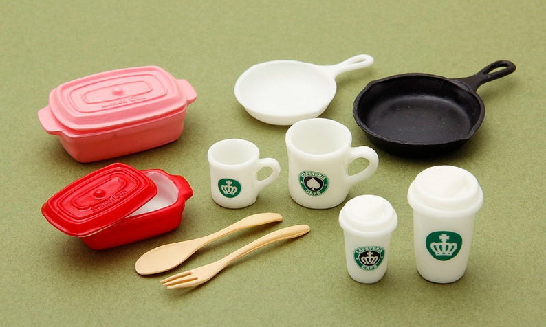 May include: A set of miniature kitchen and coffee shop items, including a pink and red casserole dish, a black skillet, a white frying pan, two white mugs with green logos, and two white to-go coffee cups with green logos. There are also two wooden utensils, a spoon and a fork.