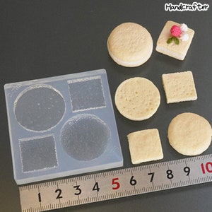 May include: A clear silicone mold with four round and two square cavities. The mold is used to create miniature cakes and cookies. There are six miniature cakes and cookies next to the mold, including a round cake with a pink and white frosting.