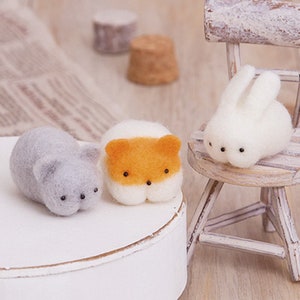 May include: Three small felt animal figurines: a grey cat, an orange and white fox, and a white rabbit. The cat and fox are lying down, while the rabbit is sitting on a small wooden chair.