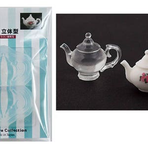 Puede incluir: Dos teteras, una transparente y otra blanca con rosas rosas, se muestran junto a un molde de plástico para hacer teteras. El embalaje dice "Clay Jewelry Die, Teapot B, 3D Shape".