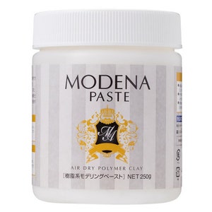 May include: A white plastic jar of Modena Paste, an air dry polymer clay. The jar has a white lid and a label with the words "MODENA PASTE" in large black letters. The label also features a gold emblem and the text "AIR DRY POLYMER CLAY" and "NET 250g".