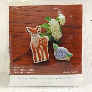 Puede incluir: Un conjunto de dos broches de fieltro bordados, uno de un cervatillo marrón y blanco manchado y el otro de una flor de hortensia azul. Los broches se muestran sobre una superficie de madera con un fondo blanco.