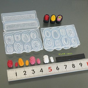 Puede incluir: Un juego de moldes de silicona transparente para hacer sushi, con una variedad de formas y tamaños. Los moldes se muestran con piezas de sushi en miniatura hechas de arcilla polimérica, incluyendo salmón, atún y atún de aleta amarilla.