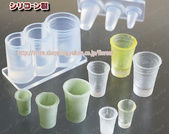 Peut inclure: Moules en silicone transparents pour le bricolage, présentant diverses formes et tailles de tasses. L'image comprend des moules avec trois tasses connectées, des tasses individuelles et des tasses en résine finies en vert et jaune. Texte japonais : "Soft Mold" et "Silicone Made."