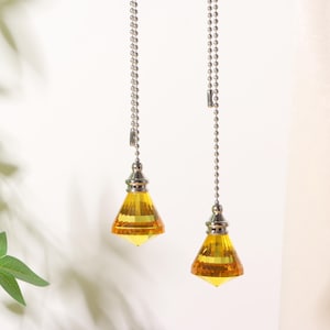 Op de afbeelding: Twee trekkoorden voor plafondventilatoren met gefacetteerde, amberkleurige kristallen hangers. Elke hanger is bevestigd aan een zilverkleurige metalen connector en een lange, dunne zilveren kogelketting voor verlichting of ventilatoren.