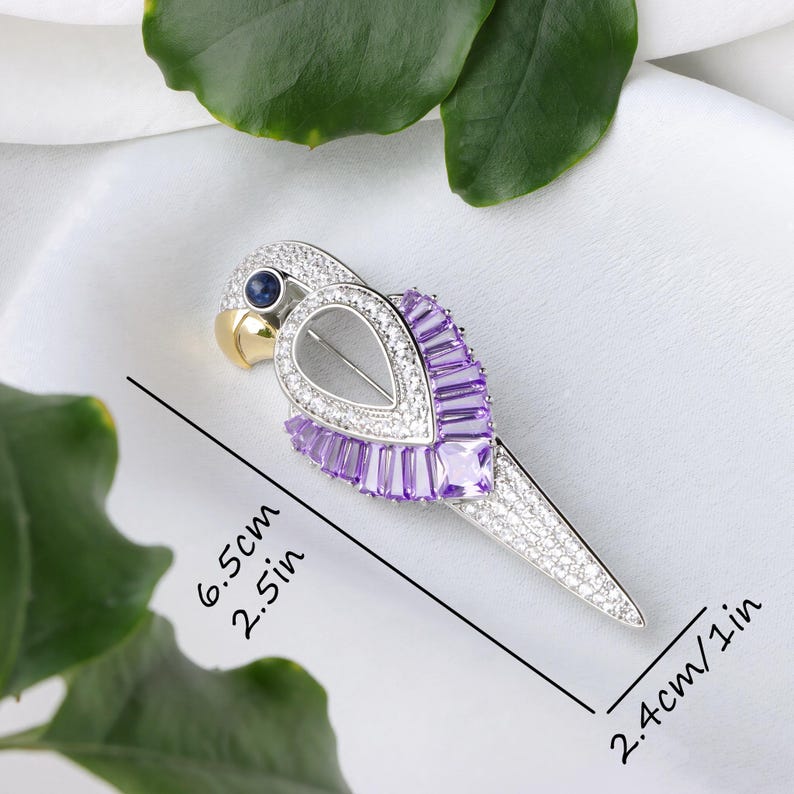 Puede incluir: Broche con forma de loro, de 6,5 cm de largo, elaborado en metal plateado, adornado con cristales transparentes y morados. El loro presenta un pico dorado, un ojo de gema azul y un cuerpo plateado. La broche se encuentra sobre un fondo blanco.