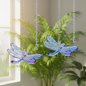 May include: Two blue dragonfly-shaped light pull chains hanging from silver ball chains. The dragonflies have intricate wing details and a gradient of blue and purple. The background features green plants and a window.