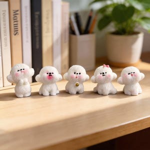 May include: Five small, white, fluffy dog figurines with pink cheeks and black eyes are arranged on a wooden surface. One has a gold bell, and another has a pink bow. The figurines are in a row, with books and a plant in the background.