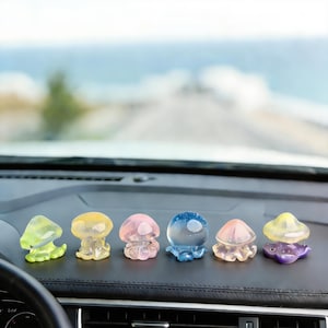May include: A collection of six colorful, translucent jellyfish figurines displayed on a car dashboard. The jellyfish are in shades of green, yellow, pink, blue, and purple. The background shows a blurred view of a road and the ocean.