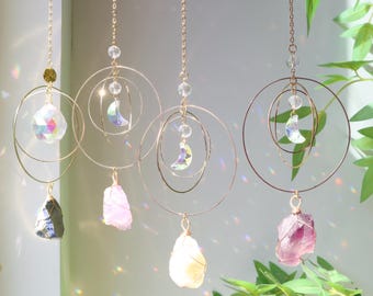 May include: A selection of hanging crystal suncatchers. Each suncatcher has a gold-coloured metal ring design with a crystal moon and a raw gemstone pendant. The gemstones are various colours, including clear, pink, and purple.