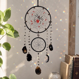 May include: A black dreamcatcher featuring two circular hoops connected by a small skull bead. The upper hoop contains a web with a central spider figure adorned with red and clear beads. Five strands of black stones and agate slices hang below.
