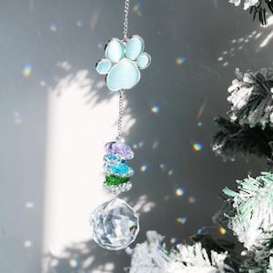 Può includere: Ornamento da appendere con un ciondolo a forma di zampa di colore azzurro chiaro, collegato tramite una catena d'argento a una pila di perline sfaccettate colorate e una sfera di cristallo trasparente. L'ornamento è progettato per catturare la luce, creando un effetto arcobaleno.