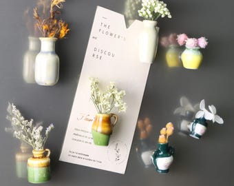 Op de afbeelding: Een verzameling kleine keramische vazen in verschillende kleuren, elk met gedroogde bloemen. De vazen zijn gerangschikt op een grijze ondergrond, met een kaart met de tekst "THE FLOWER'S and DISCOURSE".