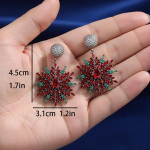 May include: A pair of snowflake-shaped earrings. Each earring features a central red gemstone surrounded by red and green accent stones. The earrings measure 1.7 inches in length and 1.2 inches in width.