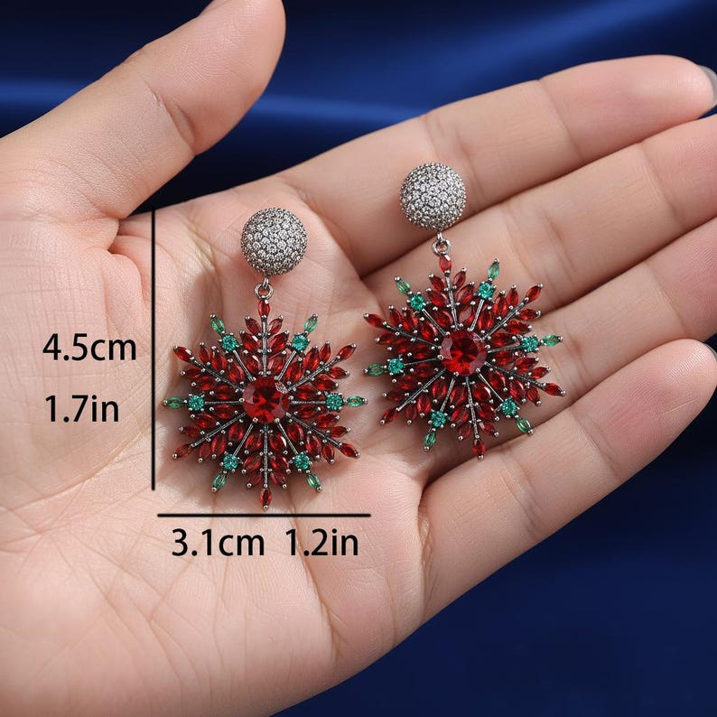 May include: A pair of snowflake-shaped earrings. Each earring features a central red gemstone surrounded by red and green accent stones. The earrings measure 1.7 inches in length and 1.2 inches in width.