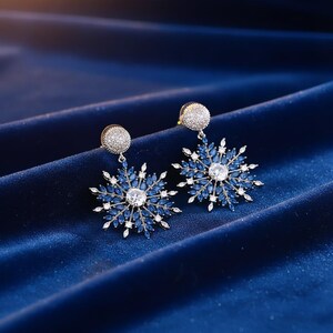 May include: A pair of snowflake-shaped earrings. Each earring features a central, clear, round stone surrounded by blue and white accent stones. The earrings have a silver-colored post and are set against a dark blue, textured background.