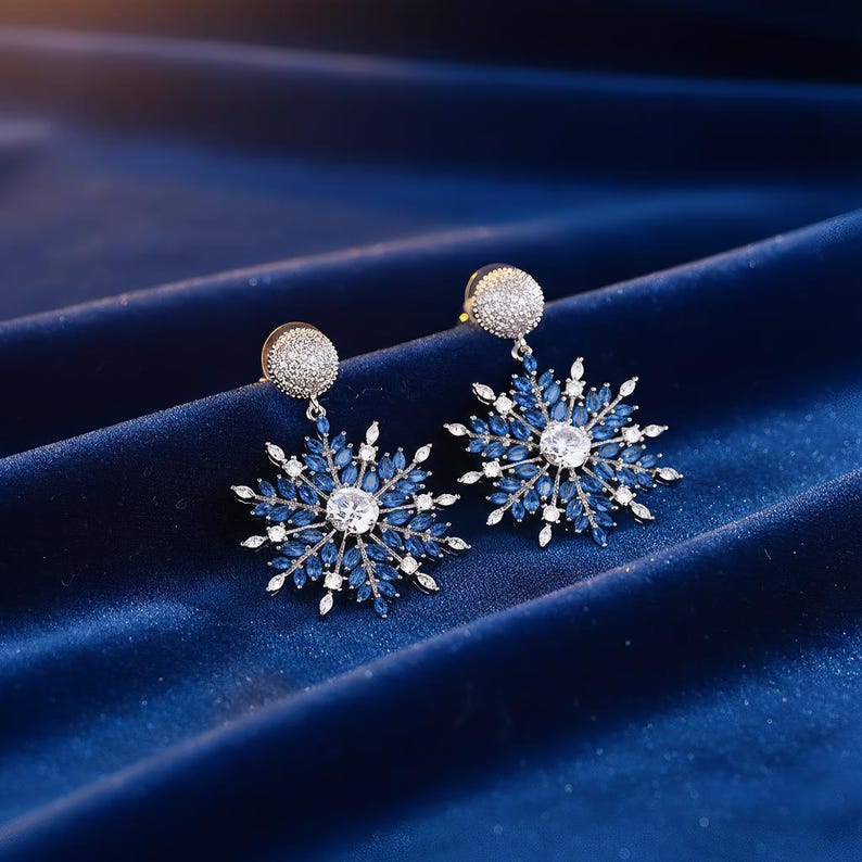 May include: A pair of snowflake-shaped earrings. Each earring features a central, clear, round stone surrounded by blue and white accent stones. The earrings have a silver-colored post and are set against a dark blue, textured background.