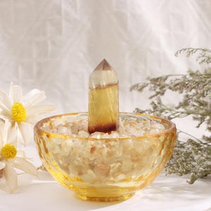 May include: A yellow and brown crystal point stands in a decorative glass bowl filled with small, light-colored stones. The bowl is a warm amber color, and the crystal has a pointed top. The background includes white flowers and dried plants.