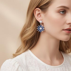 May include: Dangle earrings featuring a snowflake design. The earrings have a silver-colored post and a large, blue and white snowflake pendant. The snowflake is made of small, round blue and white stones. The earrings are worn by a person with blonde hair.