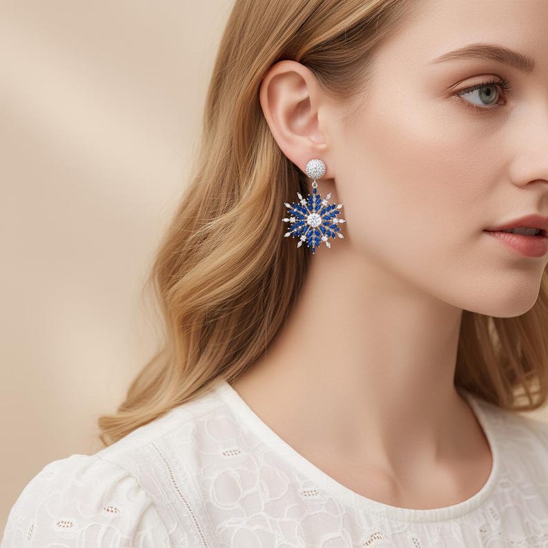 May include: Dangle earrings featuring a snowflake design. The earrings have a silver-colored post and a large, blue and white snowflake pendant. The snowflake is made of small, round blue and white stones. The earrings are worn by a person with blonde hair.