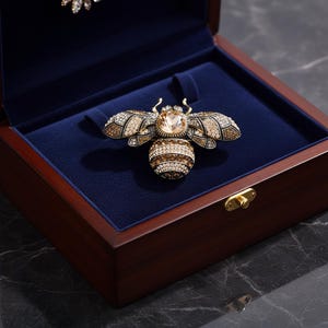 Könnte beinhalten: Goldfarbene Bienenbrosche mit einem großen, hellfarbigen Edelstein in der Mitte und Kristallakzenten auf Flügeln und Körper. Die Brosche wird in einer dunklen Holzbox mit blauem Samtinterieur präsentiert.