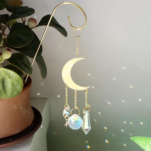 May include: A gold-coloured metal crescent moon suncatcher suspended from a curved hook. It features three crystal pendants hanging from chains. A potted plant is visible in the background.