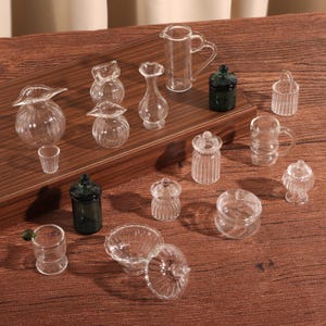 May include: A collection of miniature glass containers, including jars, vases, pitchers, and cups. The clear glass pieces are arranged on a wooden surface, showcasing their delicate shapes and designs. Some containers have lids or handles.